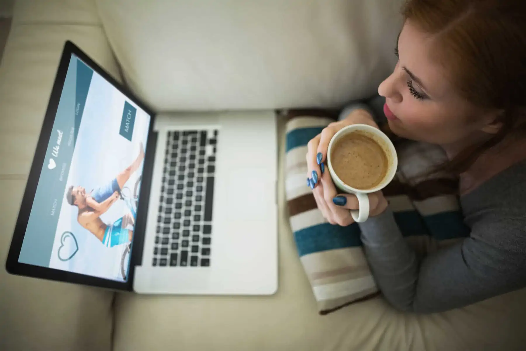 A woman sitting on a couch holding a cup of coffee in front of a laptop