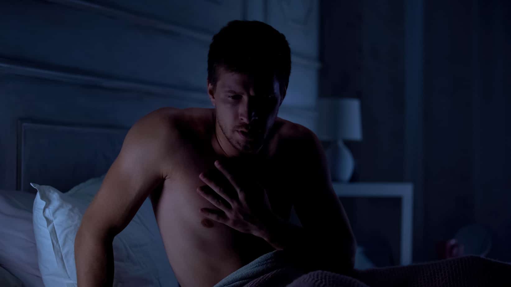 A shirtless man sitting on a bed in the dark