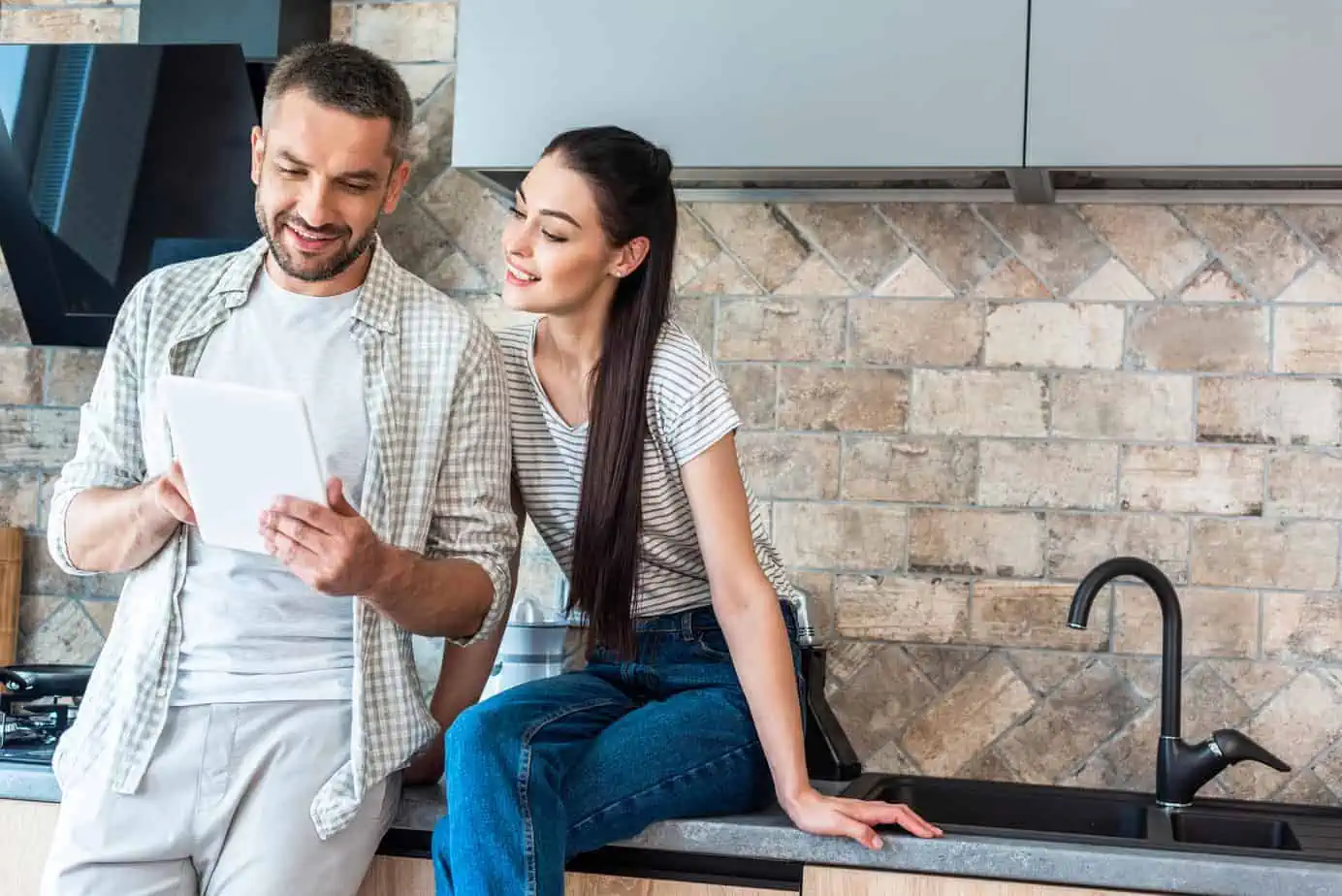 A man and woman sitting on a kitchen counter looking at a tablet