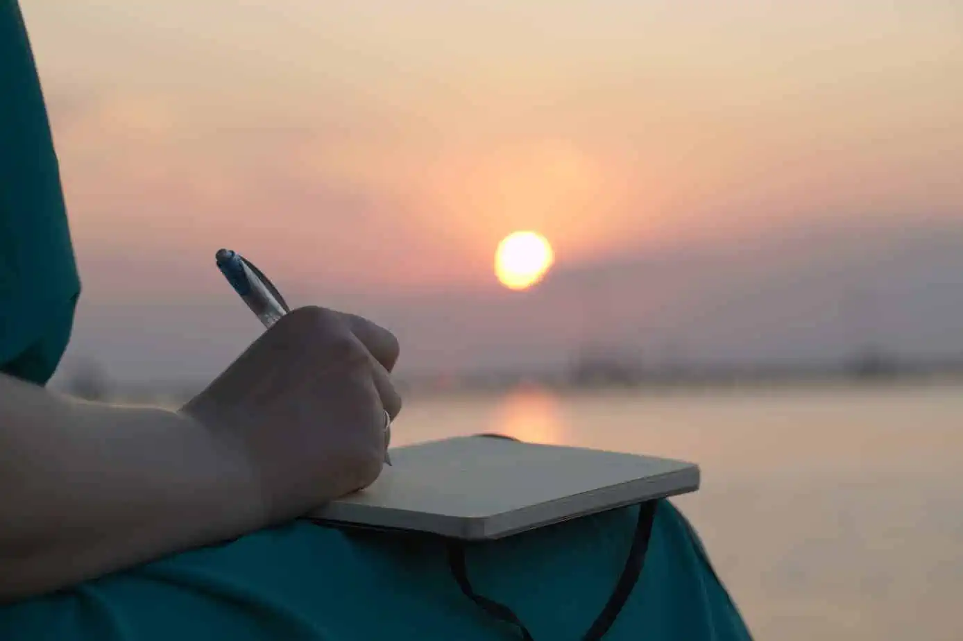 A person writing on a notebook in front of a sunset